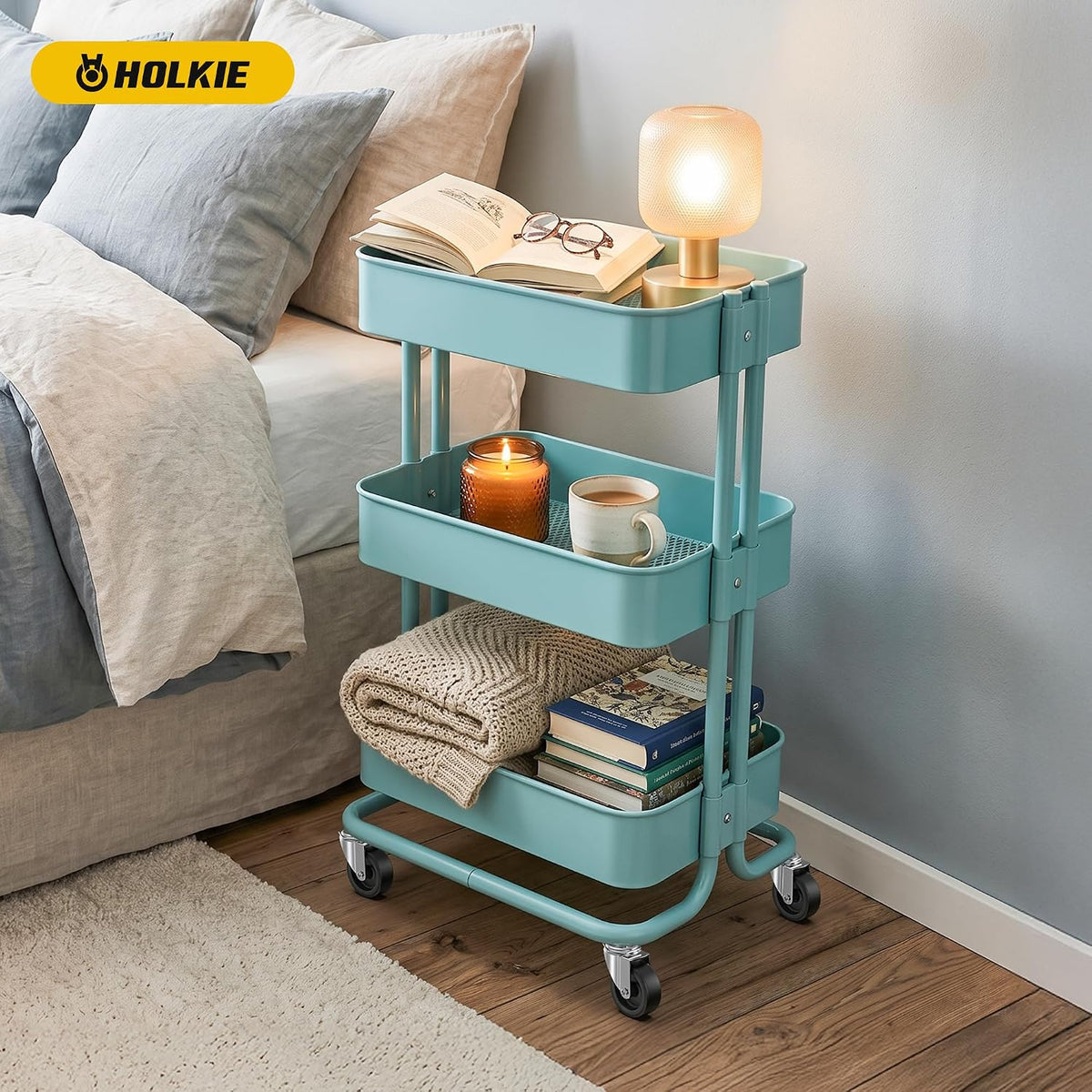 A light blue three-tier rolling storage cart styled in a bedroom corner next to a bed, holding an book, a glowing lamp, a candle, a mug, folded blankets, and books.